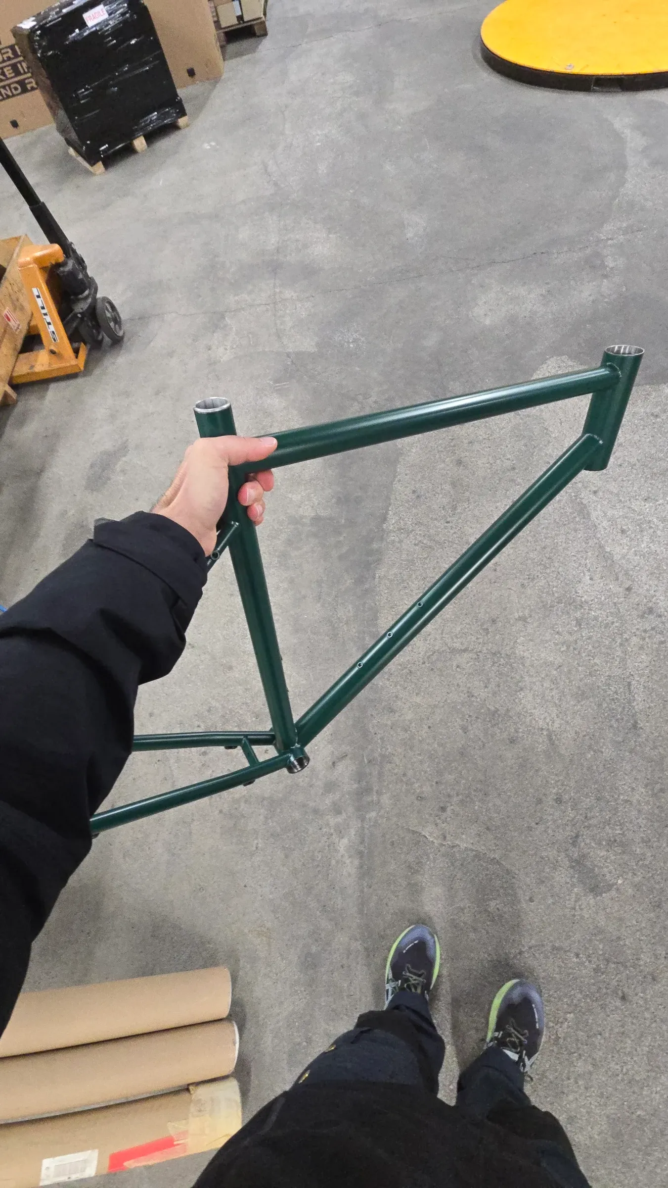 Cadre de vélo vert foncé tenu à la main dans un atelier, vue de dessus avec jambes et équipements en arrière-plan